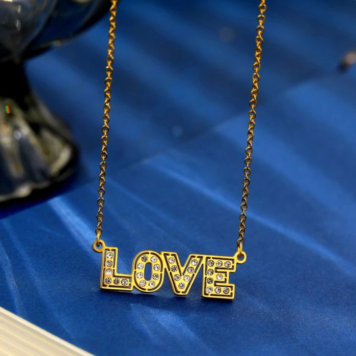 The word LOVE, in all uppercase gold letters, studded with clear crystals, suspended from a gold chain